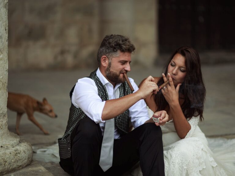 Bride and groom sitting outdoors enjoying cigars together on their wedding day.