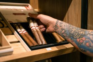 Tattooed hand holding a box of premium cigars in a wooden humidor, highlighting luxury and style.